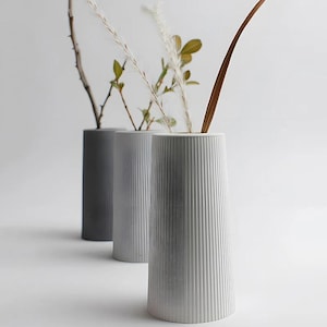 May include: Three contemporary, cylindrical vases in shades of grey. The vases feature vertical ribbed detailing and contain dried plants and foliage. The vases are arranged in a row, with the front vase in focus.