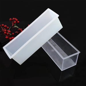 May include: Two rectangular soap molds. One is opaque white, the other is transparent. Both are empty and have a long, narrow shape. The molds are made of a flexible material, likely silicone, and are designed for soap making.