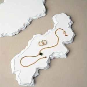Könnte beinhalten: Eine goldfarbene Halskette mit einem grünen quadratischen Anhänger, zwei goldfarbenen Ringen und eine goldfarbene Kette werden auf einer weißen, strukturierten Oberfläche präsentiert. Der Schmuck ist auf einer weißen, geschichteten, abstrakten Form vor einem neutralen Hintergrund angeordnet.