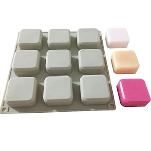 Puede incluir: Un molde de silicona gris con nueve cavidades cuadradas, cada una con una tapa a juego. Tres jabones cuadrados individuales en rosa, melocotón y rosa pálido están junto al molde. El molde está diseñado para hacer jabones cuadrados u otras manualidades.
