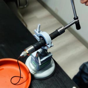 May include: A close-up of a metal tool with a black hose clamped in place. The tool is silver and gray, with a handle and a nozzle. An orange tray sits in the foreground.