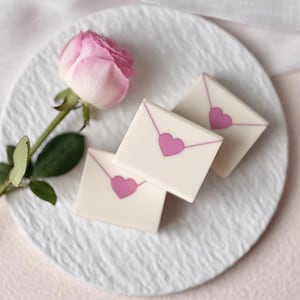 May include: Three square, off-white candies with pink heart designs and envelope outlines are arranged on a white plate. A pink rose with green leaves is next to the candies. The plate has a textured surface.