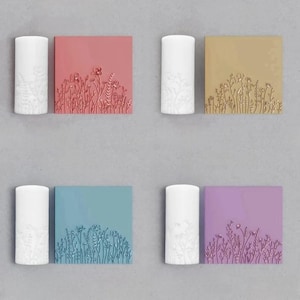 Może przedstawiać: Cztery zestawy białych świec z kwiatowymi wzorami po bokach, każda sparowana z kwadratową płytką z kwiatowym wzorem w innym kolorze: różowym, złotym, niebieskim i fioletowym.