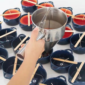 Puede incluir: Una persona vierte cera en peque&ntilde;os recipientes de cer&aacute;mica azul usando una olla de vertido de acero inoxidable con un pico de vertido de resorte. Los recipientes tienen palos de madera en la parte superior, y la cera es de color rojo oscuro.