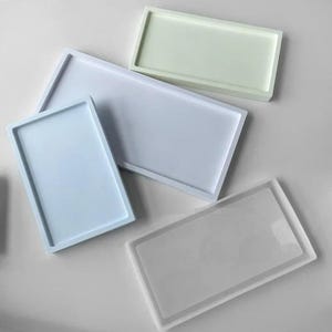 Può includere: Quattro vassoi rettangolari di varie dimensioni e colori. Un vassoio è verde pallido, uno azzurro, uno bianco e uno trasparente. Ogni vassoio presenta un bordo rialzato e una superficie interna piatta e liscia per appoggiare oggetti.