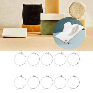 Könnte beinhalten: Eine Auswahl handgemachter Seifen in verschiedenen Formen und Farben, darunter Schwarz, Grün und Beige. Ein weißer Seifenschneider ist ebenfalls zu sehen, zusammen mit mehreren silberfarbenen Metallreifen.