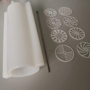 Pu&ograve; includere: Uno stampo in silicone bianco di forma cilindrica, un'asta di metallo e diversi disegni ovali bianchi tagliati al laser. I disegni presentano vari motivi geometrici e floreali. Gli articoli sono disposti su una superficie grigia.