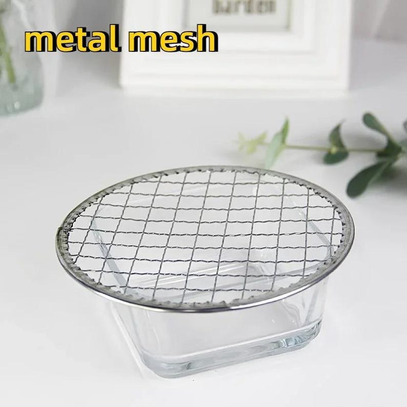 May include: A round metal mesh grill sits on top of a clear glass square bowl. The grill is made of thin metal wires and has a silver finish. The bowl is empty.