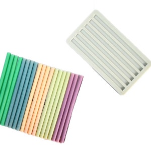 May include: An assortment of colorful glue sticks in shades of green, blue, peach, yellow, and purple, alongside a white rectangular silicone mold with parallel grooves. The items are arranged on a white background.