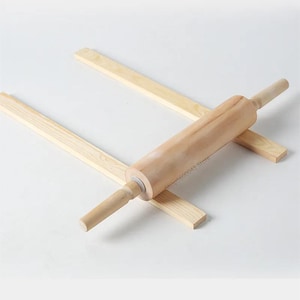 Könnte beinhalten: Ein Nudelholz aus Holz mit konischen Griffen, das auf zwei hellfarbenen Holzstützen ruht. Das Nudelholz ist aus hellfarbenem Holz gefertigt und für das Backen und Kochen bestimmt. Das Bild ist vor einem weißen Hintergrund aufgenommen.