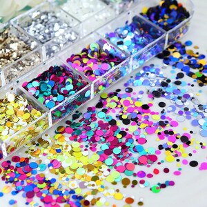 May include: A clear plastic organizer filled with colorful, round sequins. The sequins are in various colors, including gold, silver, pink, blue, and green. Some sequins are scattered outside the organizer. Ideal for nail art, crafts, or other decorative projects.