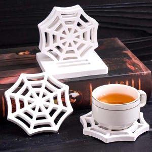 Puede incluir: Posavasos blancos con forma de telaraña. Un posavasos sostiene una taza de té blanca con té. Dos posavasos están apilados y otro se coloca por separado. Los posavasos están hechos de un material blanco sólido y están diseñados con un patrón de telaraña.