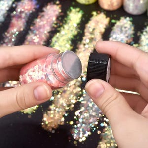 May include: A small glass jar filled with pink and iridescent glitter, with the black lid removed. The glitter is scattered on a surface, with other jars of glitter in the background. The jar is being held by a person.