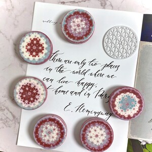 Pu&ograve; includere: Vista dall'alto di sette oggetti decorativi rotondi con intricati motivi geometrici nei toni del bordeaux, bianco e azzurro. Sullo sfondo, un foglio bianco con testo scritto a mano. Citazione di Hemingway.