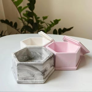 Op de afbeelding: Drie zeshoekige betonnen dozen in wit, roze en gemarmerd grijs. De roze doos heeft het deksel iets geopend. De dozen staan op een wit oppervlak, met groen gebladerte op de achtergrond. Deze decoratieve dozen zijn ideaal voor kleine items.