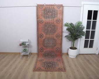 Puede incluir: Una alfombra larga y rectangular de aspecto desgastado, con una combinación de colores rojo y marrón descoloridos. La alfombra presenta un patrón geométrico repetitivo con motivos florales y de medallones. Se muestra contra una pared neutra, con una puerta blanca y una planta en maceta en el fondo.