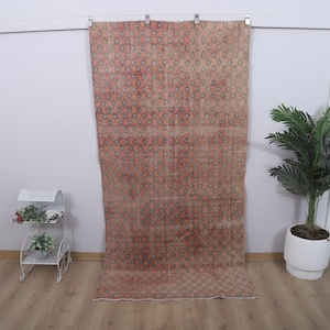 May include: A vintage, rectangular rug with a faded, patterned design in shades of red, brown, and green. The rug is hanging against a white wall. A white plant stand with flowers and a potted plant are in the foreground.