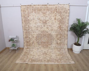 May include: A large, beige and brown patterned rug with a central medallion design. The rug is displayed against a white wall, with a small white plant stand and a potted plant to the side. The rug's dimensions are not visible.