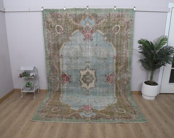 May include: A large, rectangular rug with an ornate, distressed design. The rug features a central light blue field with floral and geometric patterns, framed by a border of brown and green. The rug is displayed against a white wall.