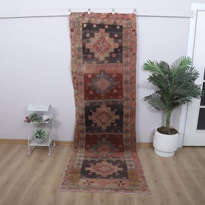 May include: A long, rectangular rug with a geometric pattern in shades of red, black, and beige. The rug is hanging on a wall, showcasing its design. A small white plant stand and a potted plant are also visible.