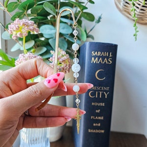 Puede incluir: Un marcapáginas dorado con una cadena de cuentas de cristal blanco y un dije de espada. El marcapáginas está siendo sostenido por una mano con esmalte de uñas rosa. El marcapáginas está frente a un libro titulado "Crescent City House of Flame and Shadow" de Sarah J. Maas.