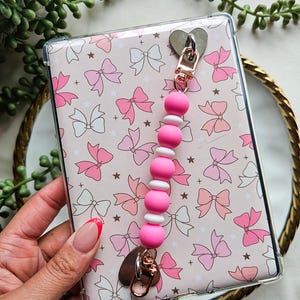 May include: A clear phone case with a pink bow pattern and a pink and white beaded wrist strap. The strap has a heart-shaped metal clasp. The case is held by a person with a French manicure.