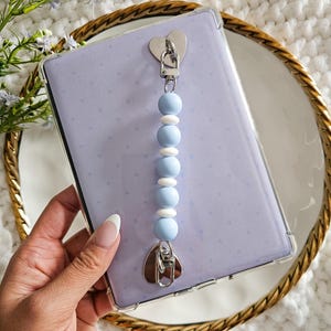 May include: A light purple phone case with a white and blue beaded phone strap. The strap is attached to the case with a silver heart-shaped charm.