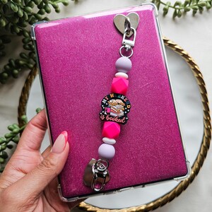 May include: A pink glittery e-reader case with a clear protective cover. Attached is a beaded charm with pink, white, and lavender beads, and a charm that says "My Weekend is Booked". The charm is attached to the case with a silver heart-shaped clip.