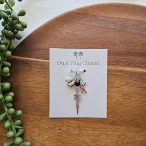 Könnte beinhalten: Eine weiße Karte mit dem Text "Dust Plug Charm" und einem silbernen Anhänger mit einem Schwert, einer schwarzen Perle, einem Buchstaben K und einem Buch-Anhänger.