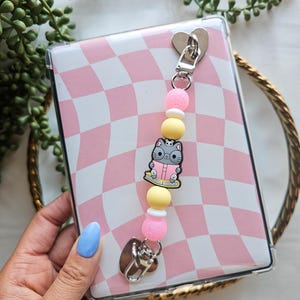 May include: A clear phone case with a pink and white checkered pattern. A gray cat charm with a book is attached to the case with a beaded chain.
