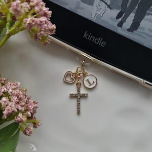May include: A gold-colored charm with a cross, heart, and initial "L" hangs from a Kindle. The cross is encrusted with small, clear crystals. The Kindle screen displays text and an illustration of a dog.