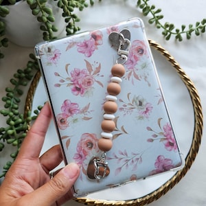May include: Clear phone case with a floral design in pink and peach tones. The case has a beaded charm with wooden and white beads, and a heart-shaped metal clasp. The case is held over a gold-rimmed mirror.