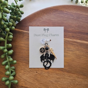 Puede incluir: Encanto de dragón negro con detalles dorados, incluyendo una espada, un cristal y un encanto con la letra "N". El encanto está en una tarjeta blanca con el texto "Dust Plug Charm".