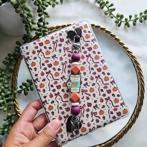 May include: A clear phone case with a fall-themed pattern of pumpkins, leaves, and ribbons. A beaded charm with a coffee cup design and the words "Caffeine Kindness" is attached. The charm features various colored beads and a heart-shaped clasp.