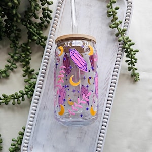 Puede incluir: Un vaso de vidrio transparente con tapa de bambú y pajita transparente. El vaso está decorado con cristales rosas, lunas crecientes, estrellas y ramitas. El vaso está sobre una bandeja de madera blanca con borde de cuentas, con plantas verdes en el fondo.