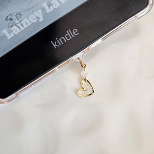 Puede incluir: Una funda transparente para teléfono con un dije de corazón dorado colgando. La funda está en un lector electrónico Kindle con el texto "Lainey Lawson" y "kindle" visible.