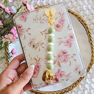 May include: A clear phone case with a floral design and a green and white beaded phone strap with gold heart-shaped clasps.