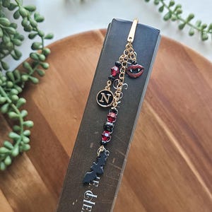 May include: A gold-colored bookmark with a chain and charms. Charms include a red-lipped vampire mouth, a black bat, and a black circle with the letter "N". Red and black beads are also attached.