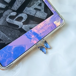 May include: A close-up of a blue butterfly-shaped charm with rhinestones. The charm is attached to a clear phone case with a holographic design.