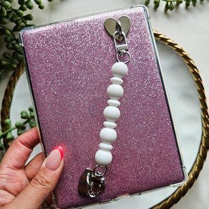 May include: A clear tablet case with a pink glitter back. A white beaded charm with a heart-shaped clasp is attached. The tablet case is on a gold-rimmed tray, with green foliage in the background.