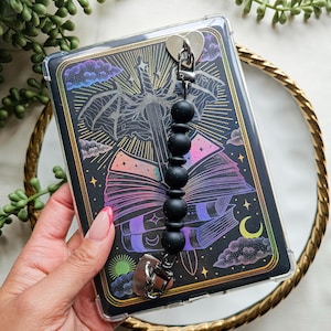May include: A clear phone case with a black graphic design featuring a bat, books, and celestial elements. The case has a black beaded strap with heart-shaped clasps. The design incorporates vibrant colors like pink, purple, and yellow.