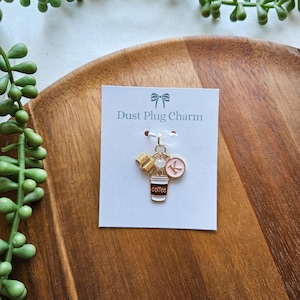 May include: A gold-toned dust plug charm with a coffee cup, book, and initial "K" pendant. The charm is attached to a white card with the text "Dust Plug Charm" in a light blue font. The background is a wooden tray.