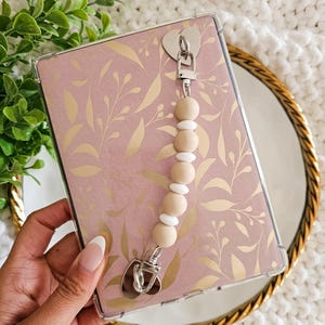 Puede incluir: Una funda transparente para teléfono con un diseño floral rosa y dorado. Una correa de teléfono con cuentas de color beige y blanco está unida a la funda.