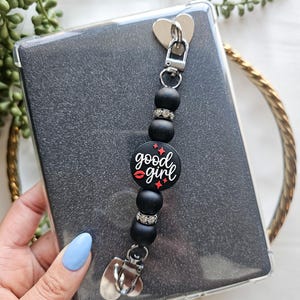 May include: Black phone charm with a round black bead with the text "good girl" in red and white lettering and a red lip graphic. The charm is attached to a clear phone case with a silver heart-shaped clip.
