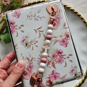 May include: A clear phone case with a floral design, featuring pink and white flowers and green leaves. A beaded charm with a cross and heart-shaped clasp is attached. The charm includes pink, white, and beige beads.