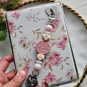 May include: A clear phone case with a floral design featuring pink and white flowers. A pink charm with the words "Faith Over Fear" is attached to the case with a beaded lanyard. The lanyard includes pink beads and wooden accents.