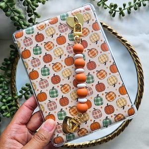 May include: A clear phone case with a pumpkin pattern, featuring orange, green, and plaid pumpkins on a beige background. The case has a gold heart-shaped charm and a beaded lanyard with orange and white beads. The case is held in a person's hand.