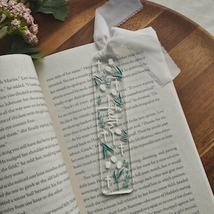 May include: A clear acrylic bookmark with white floral and green leaf accents, and the word "Bloom" in white script. A white ribbon is attached to the top. The bookmark rests on the pages of an open book.
