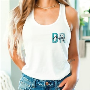 May include: White tank top with the text "DR Ashley" in teal and black script. The tank top is sleeveless and made of a soft material. The text is centered on the chest.