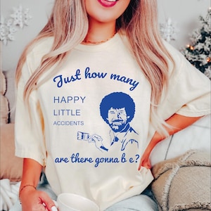 May include: A white t-shirt with a blue graphic of Bob Ross painting. The text on the shirt reads "Just how many happy little accidents are there gonna be?"